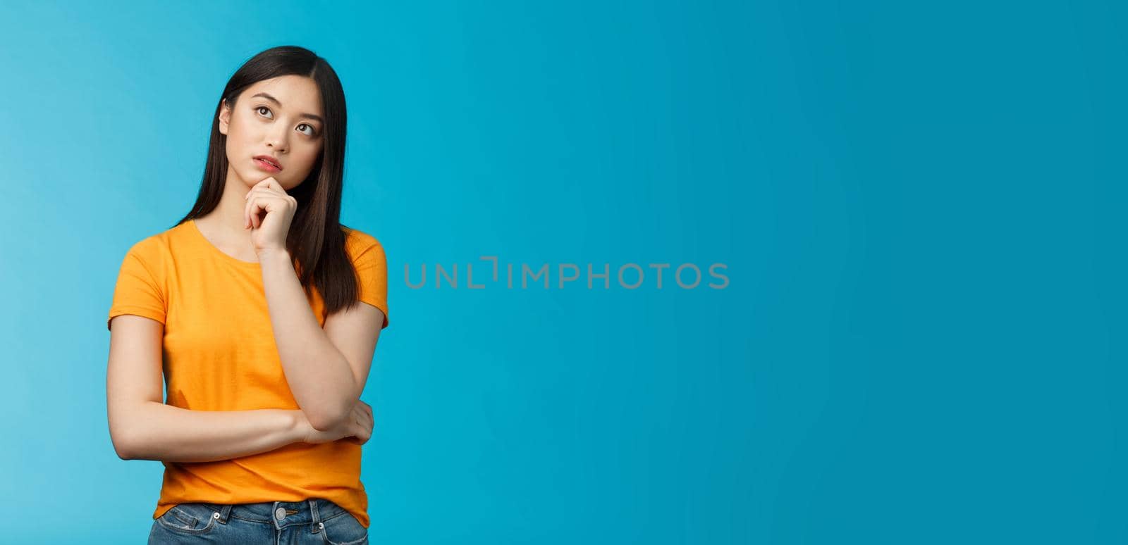 Thoughtful dreamy smart asian girl thinking, making plan inside head, touch chin, look up focused, contemplating interesting scene, pondering, making choice, taking decision, stand blue background.