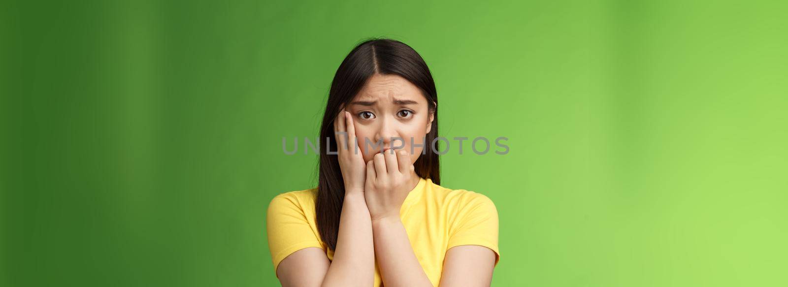 Innocent insecure timid asian scared girl panicking, standing afraid victim terrified, touch cheek shocked, frowning stunned, biting fingernails, anxiously stare camera, green background.