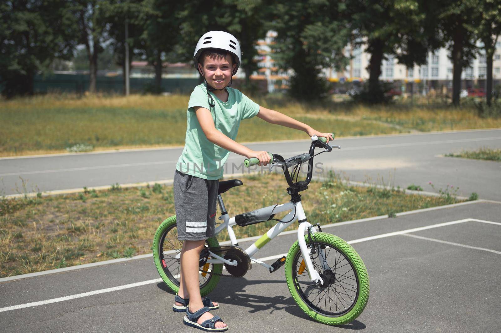 Charming active Caucasian child boy, in protective helmet, looking at camera, standing near his bicycle on the asphalt road outdoor. Physical activity. Active lifestyle. Equilibre and balance. Sport