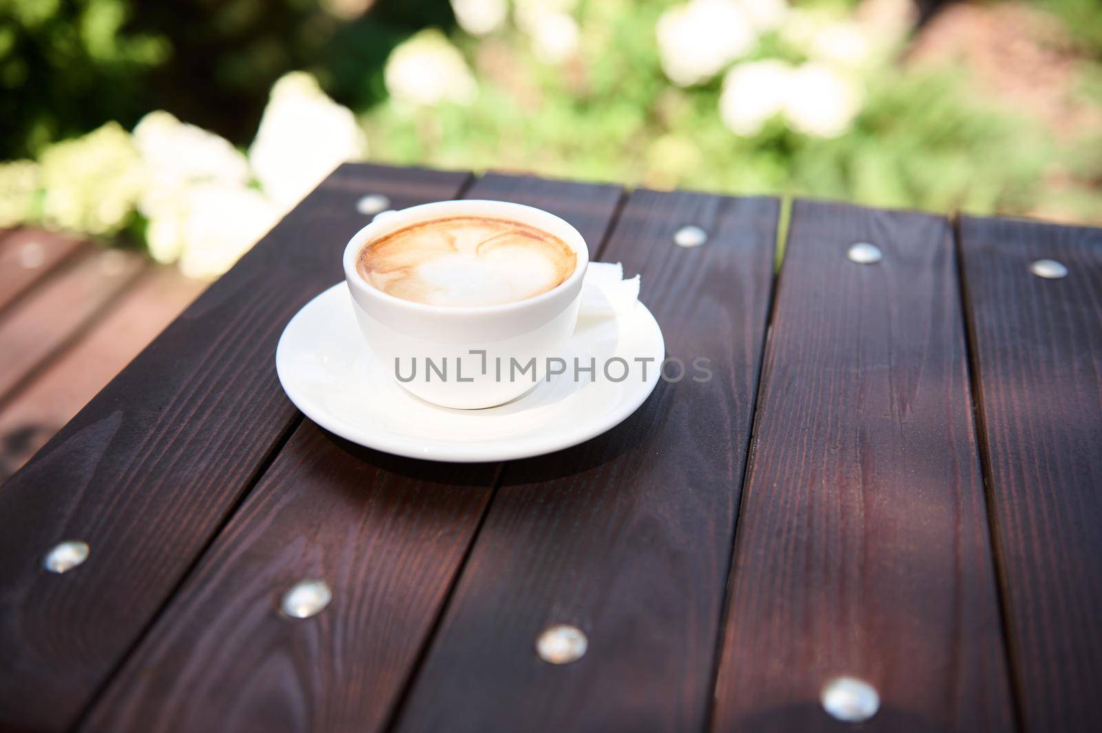 Still life with a white cup of frothy cappuccino with foam on a wooden table, in the summer terrace of a cafeteria. Food and drink