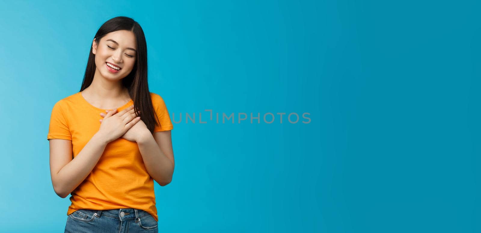 Lovely asian woman with dark haircut close eyes press hands heart gently smiling, tilt head cute cherish heartwarming memories, daydreaming, imaging beautiful dream, stand blue background.