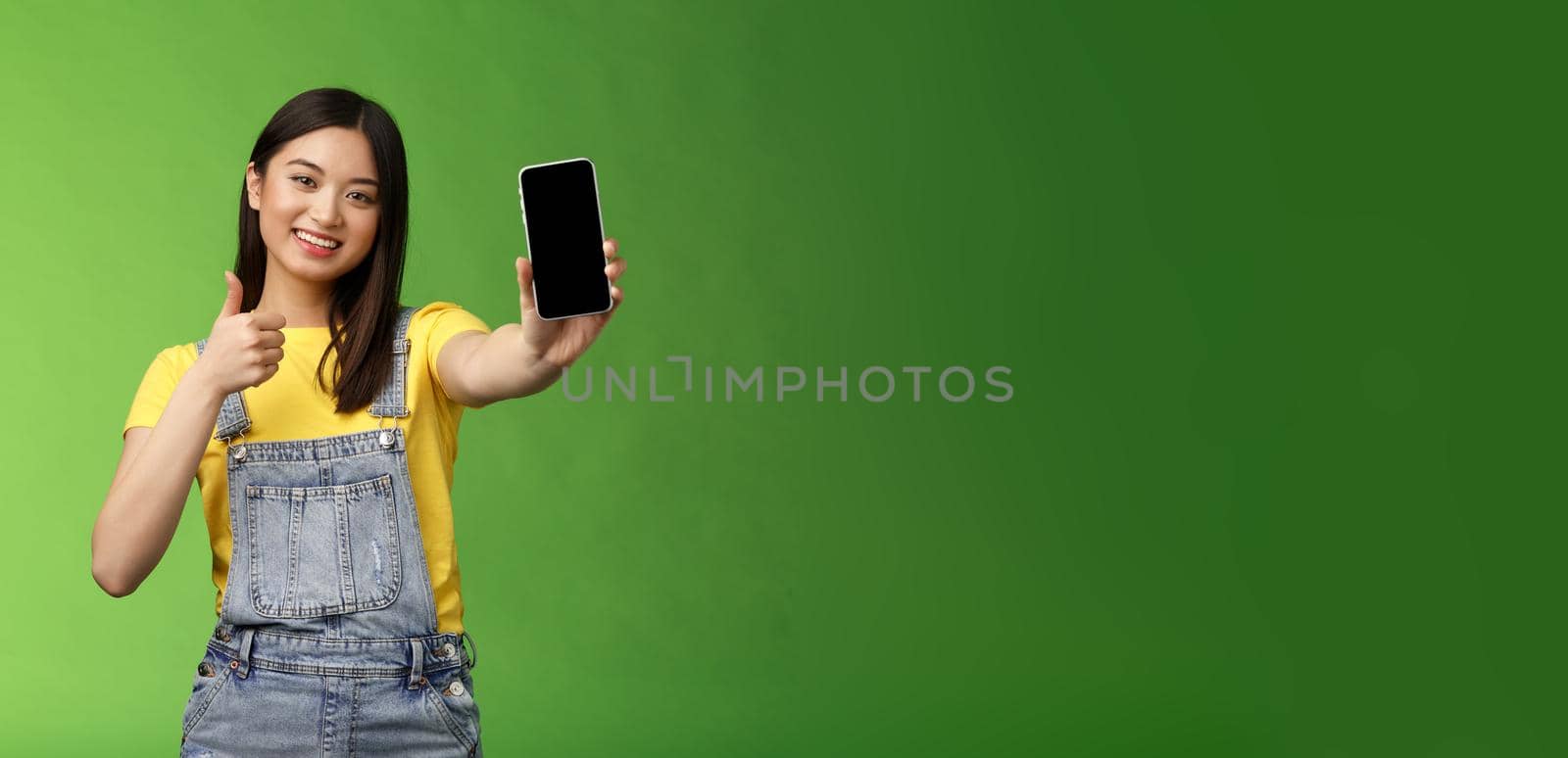 Cheerful cute asian woman judging good smartphone game, show thumb-up like sign, extend arm hold telephone screen, present awesome app, promoting application, stand green background.