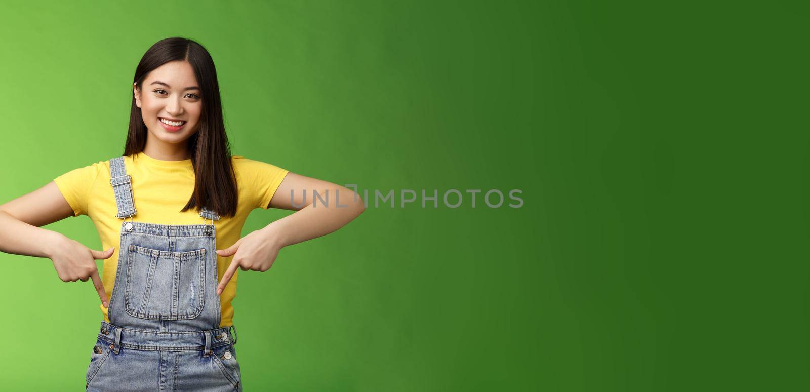friendly outgoing cute asian young woman show fingers down, introduce new promo app, smiling joyfully sharing link, recommend product using brand, grinning toothy, green background. Copy space