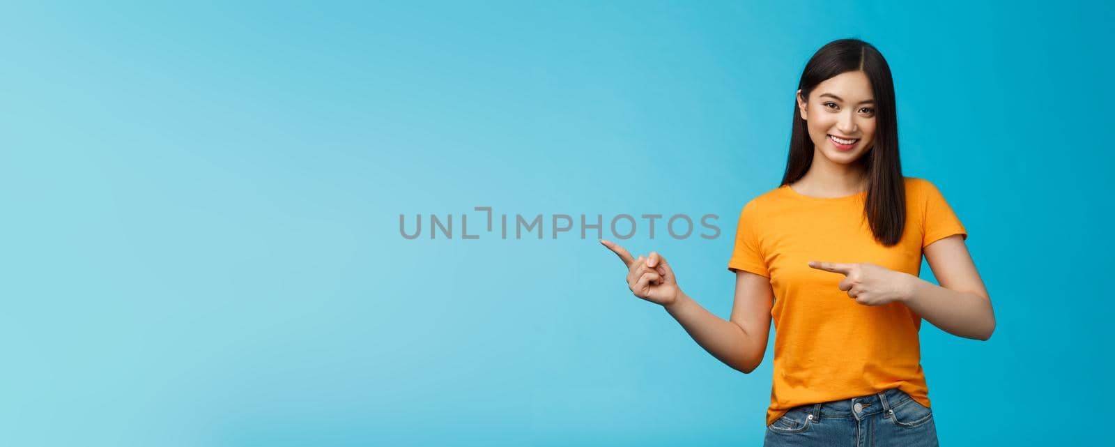 Friendly outgoing good-looking asian woman showing promo, pointing left indicating fingers advertisement, give advice, smiling confident, pleasantly help pick clothes, stand blue background.