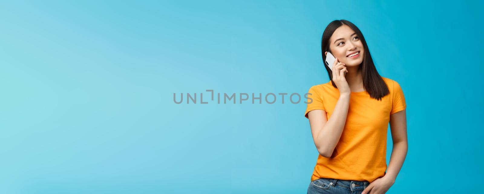 Friendly cute urban asian girl look up dreamy relaxed, smiling joyfully hold smartphone near ear, talking casually friend via telephone, satisfied good mobile connection, stand blue background by Benzoix