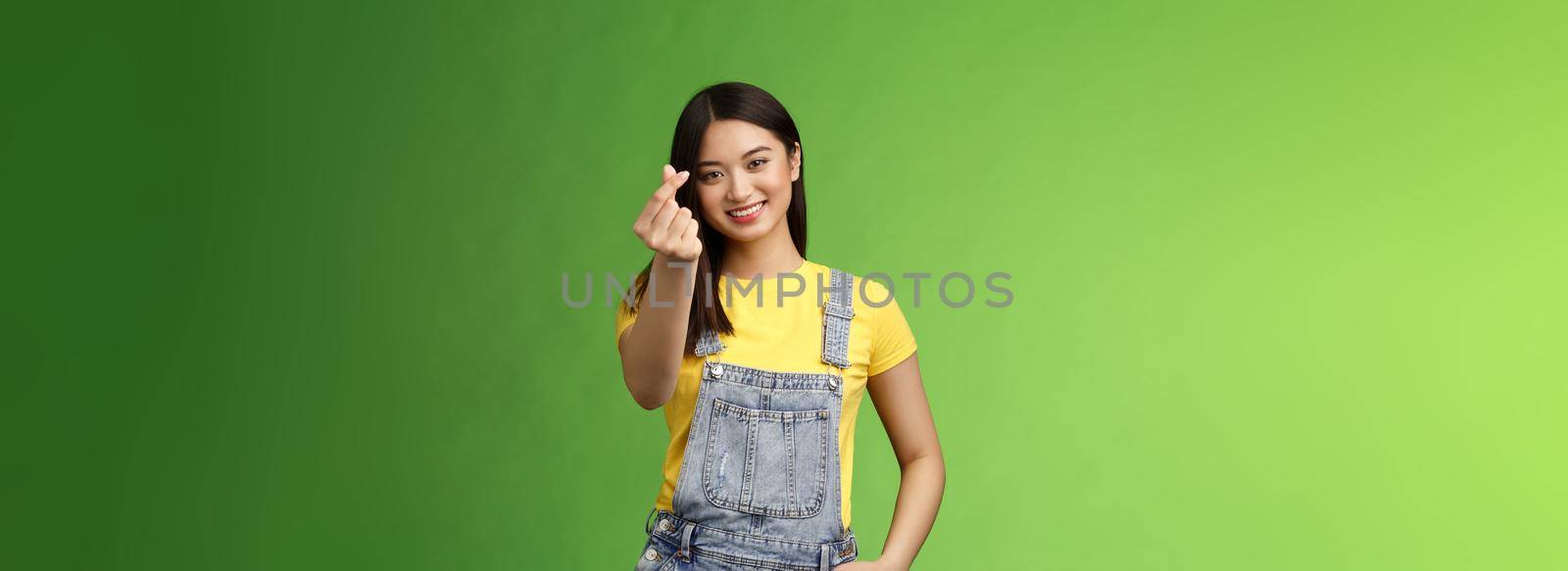 Lovely tender carefree asian girlfriend show korean love sign, make finger heart smiling cute, showing passion and affection, stand green background in overalls and yellow t-shirt, express sympathy by Benzoix