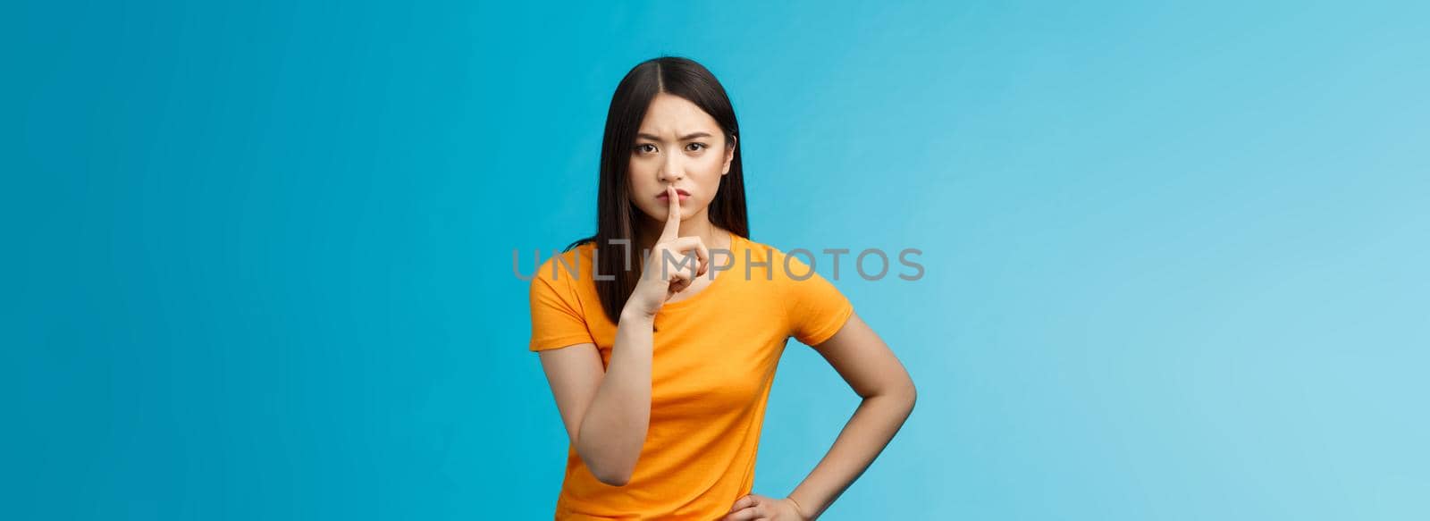 Hush keep quiet, behave well. Serious-looking strict asian elder sister shushing bending towards camera frowning, hold hand waist encouraged, asking not share secret, stand blue background.