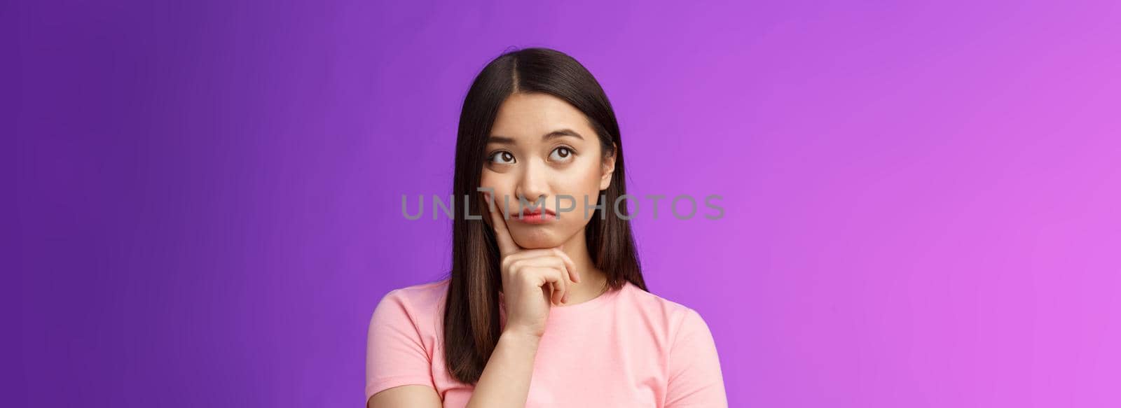 Creative dreamy asian female thinking, have tough decision mind, touch cheek thoughtful, look up, pondering making choice, stand hesitant sighing troublesome thoughts, stand purple background.