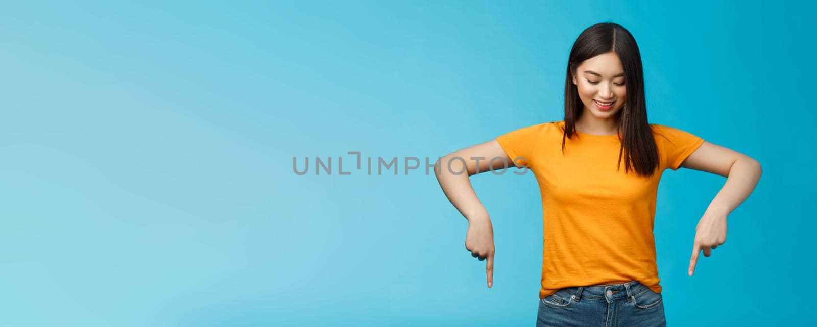 Cute tender feminine asian girl dark haircut look pointing down contemplating interesting product, stare curiously smiling amused, express enthusiasm interest try-out cool thing, blue background.