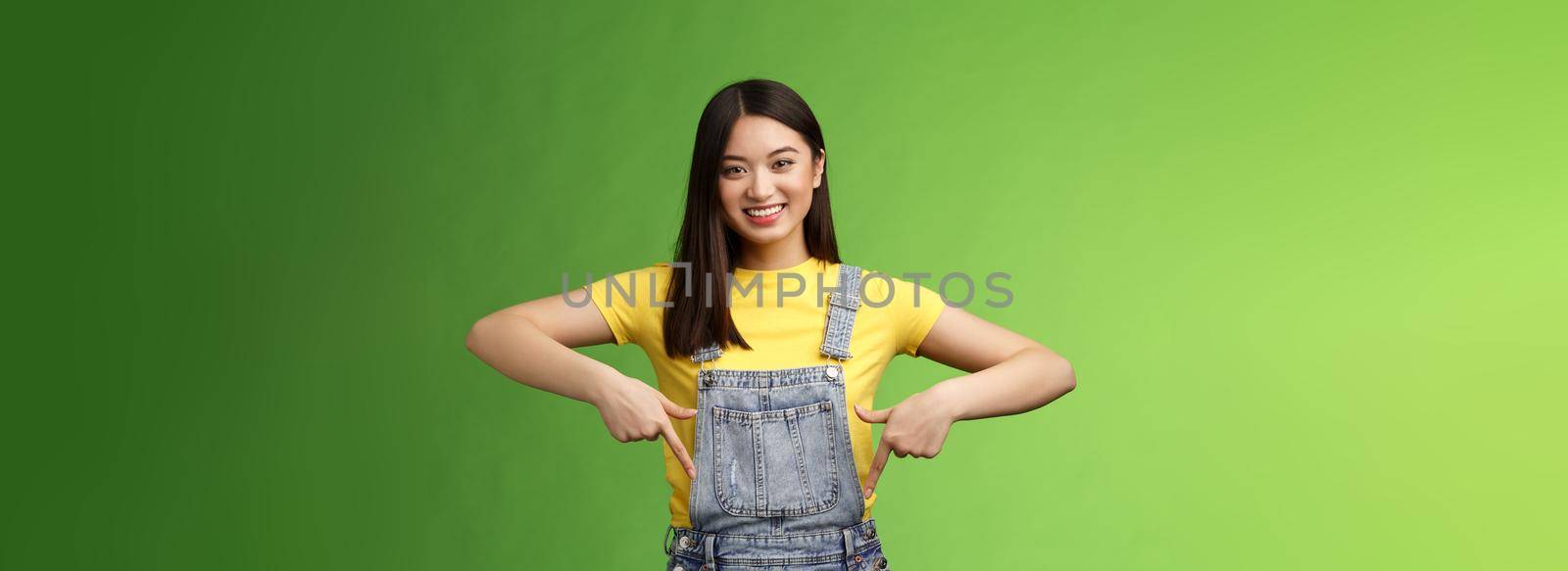 friendly outgoing cute asian young woman show fingers down, introduce new promo app, smiling joyfully sharing link, recommend product using brand, grinning toothy, green background. Copy space