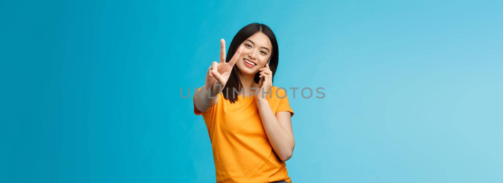 Charismatic cute girl share positive news talking smartphone show peace, victory sign smiling satisfied, standing blue background receive pleasant phone call, achieve deal via phone-call.