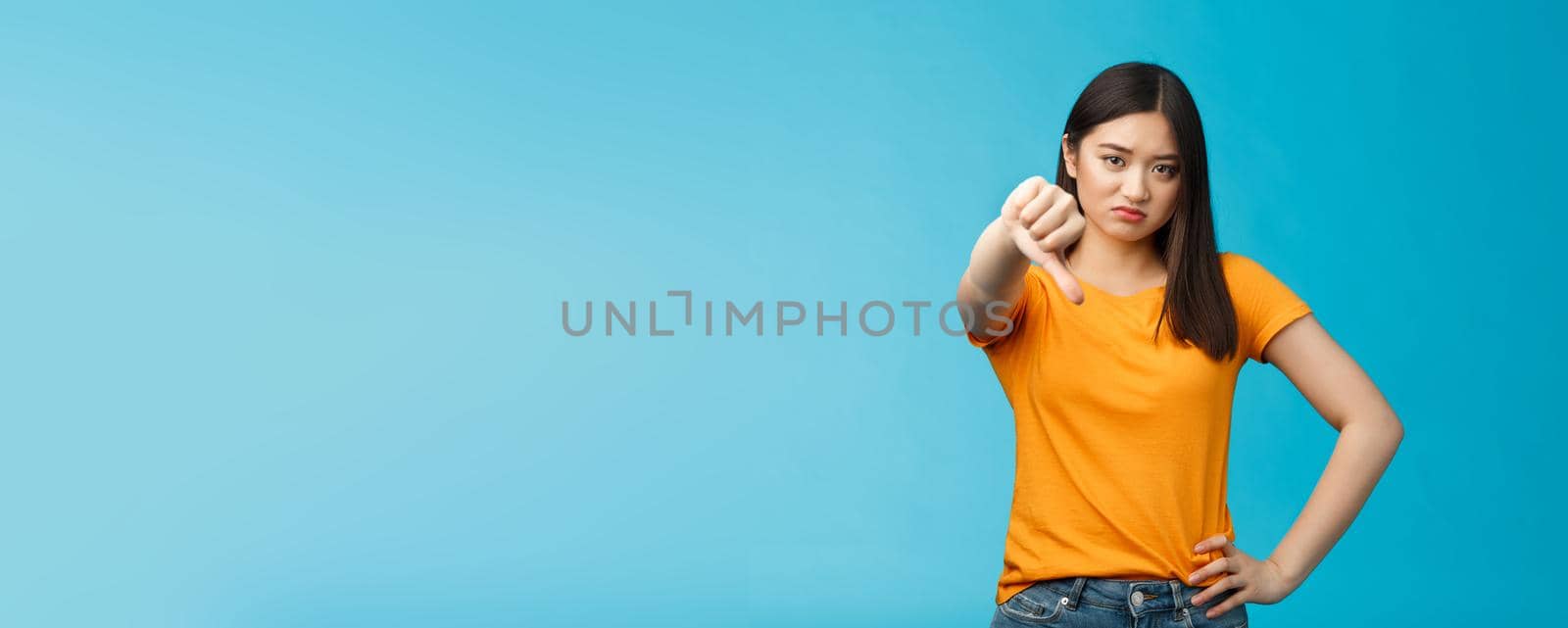 Disappointed and unsatisfied cute asian girl grimacing, showing thumb-down no, dislike sign, frowning, cringing from aversion and reluctance, stand displeased blue background by Benzoix