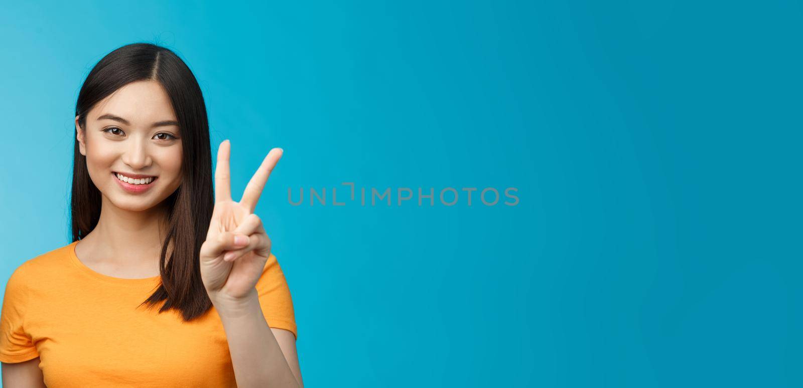 Cheerful friendly asian woman in yellow t-shirt smiling broadly, show number two, twice, win second place, smiling joyfully, make take-away order, book sits, stand blue background.