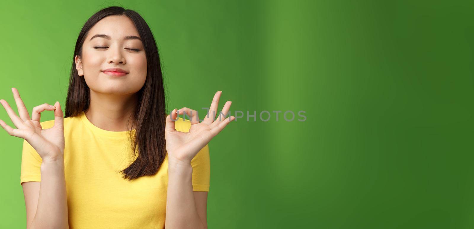 Peaceful charming asian girl brunette inhale fresh air, relaxing breathing happily, close eyes smiling, hold hands zen mudra signs reach nirvana, meditation, practice yoga, green background.
