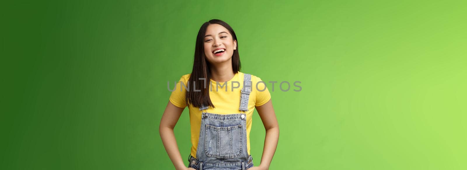 Carefree enthsuastic outgoing attractive teenage asian girl having fun laughing friends, hold hands overalls pockets smiling toothy, take party easygoing conversation, stay positive green background.