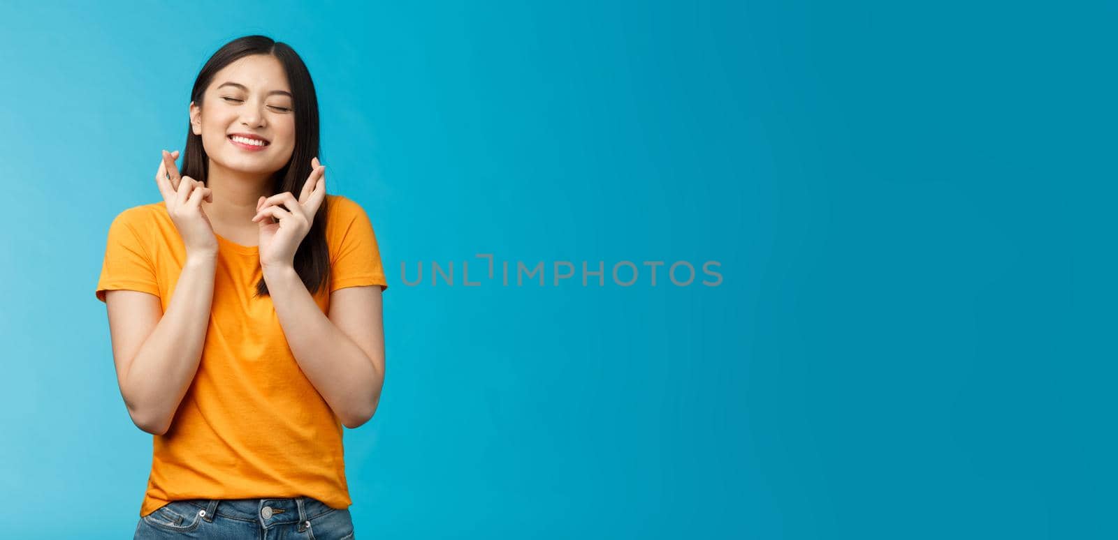 Hopeful excited asian young girl making wish cross fingers good luck, close eyes smiling believe dream come true, anticipating important event, praying good fortune, stand blue background.