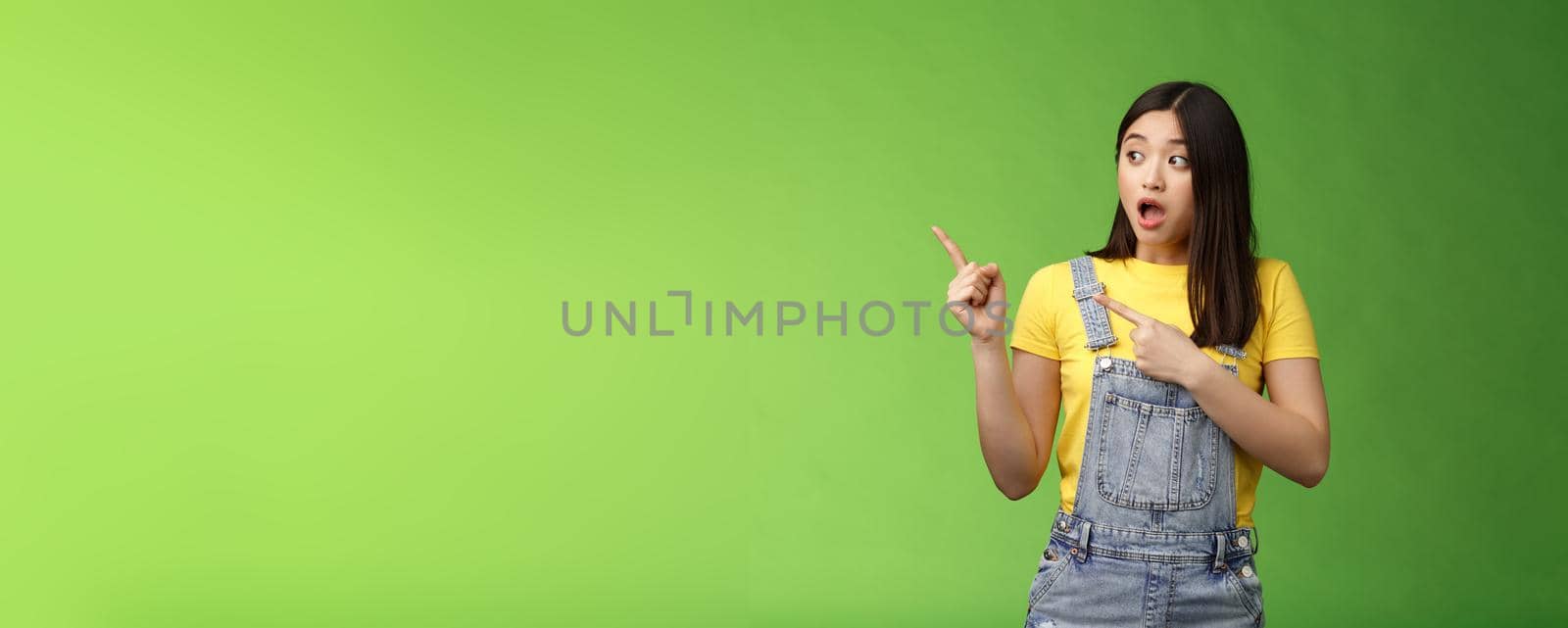 Shocked gasping alarmed pretty asian girl drop jaw impressed, turn pointing left hear loud bang, stand astonished speechless, gazing curious event, pose green background by Benzoix