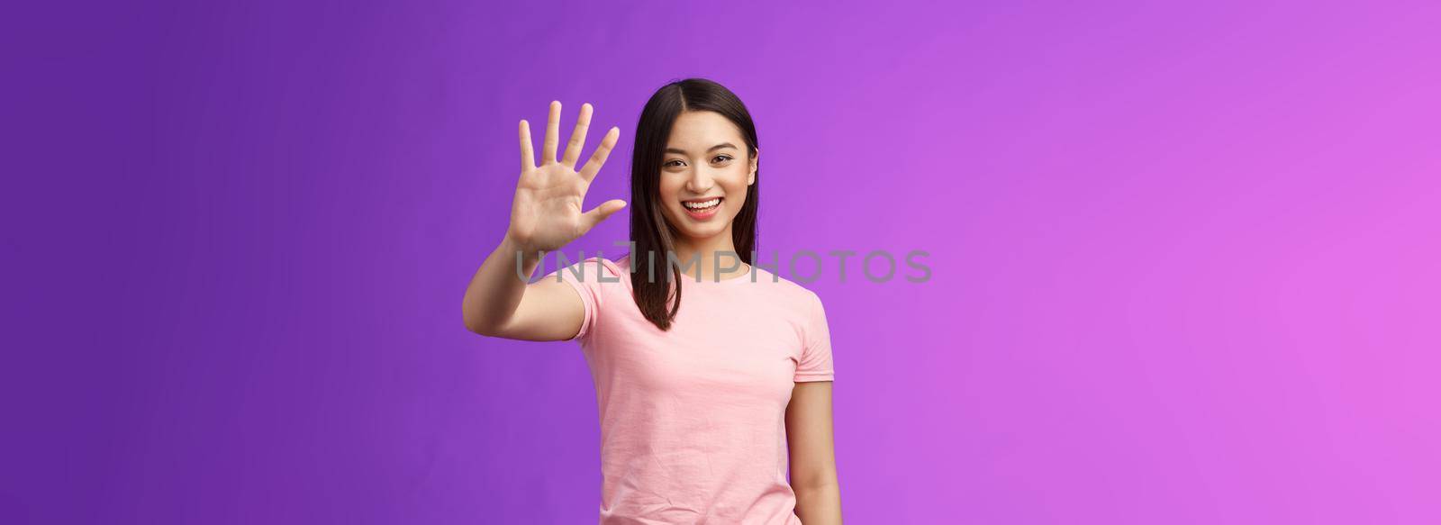 Friendly cute asian tender girl raise palm waving goodbye, smiling joyfully, farewell, gladly welcome friends inviting come in, saying hi hello, greeting, stand purple background. Copy space