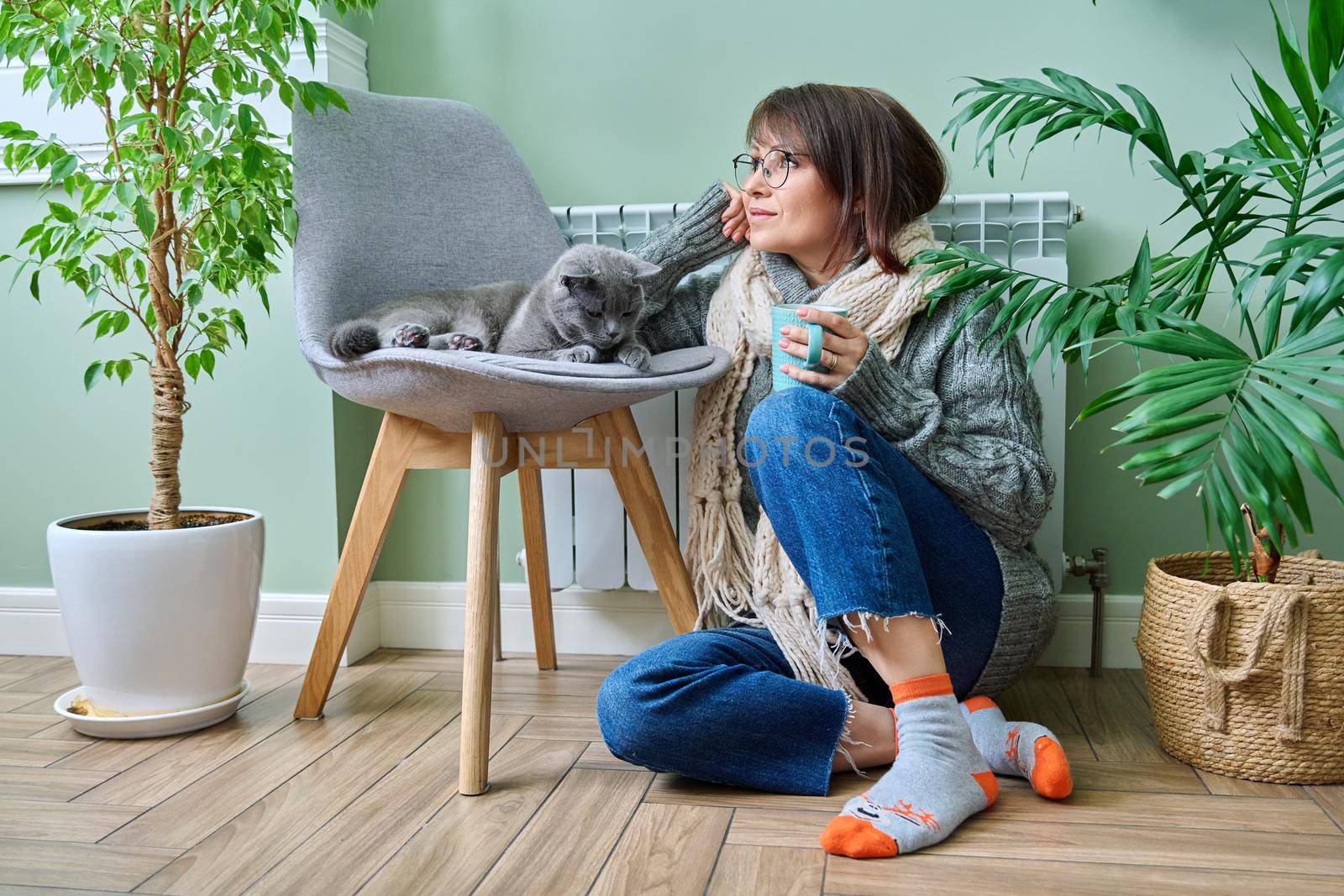 Cold winter season woman with cat near heating radiator. Middle-aged female in warm woolen knitted sweater with cup of hot drink and pet lying on chair. Heating season, lifestyle, crisis energy saving