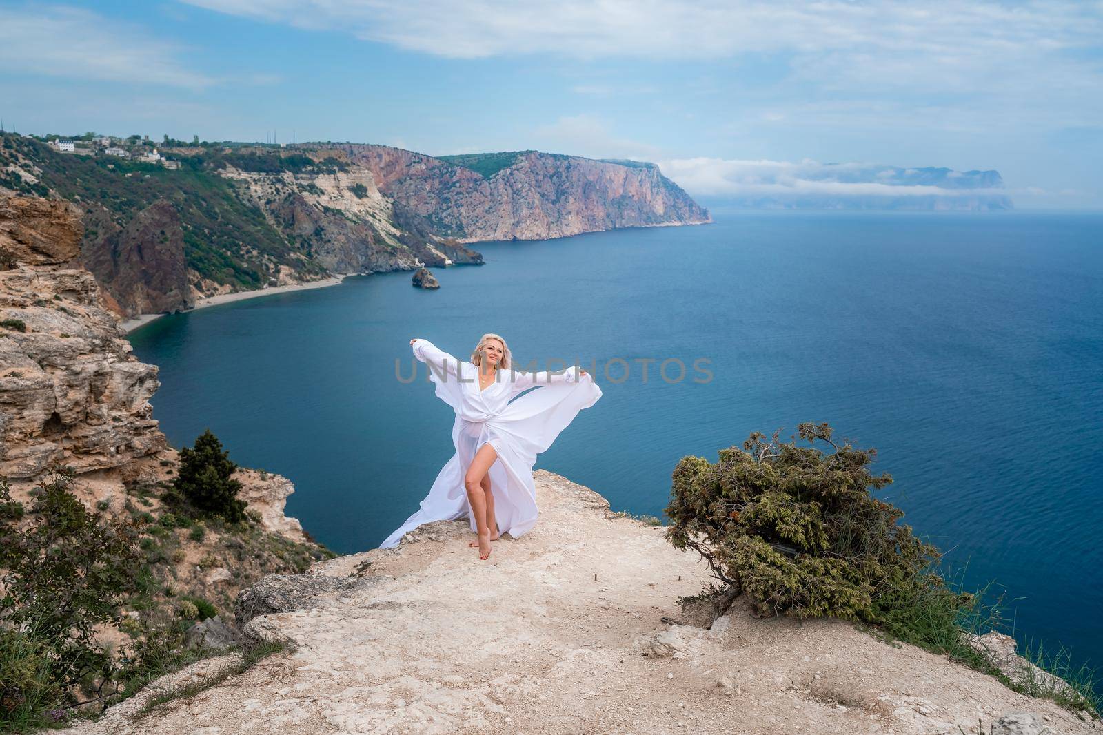 Blonde with long hair on a sunny seashore in a white flowing dress, rear view, silk fabric waving in the wind. Against the backdrop of the blue sky and mountains on the seashore. by Matiunina