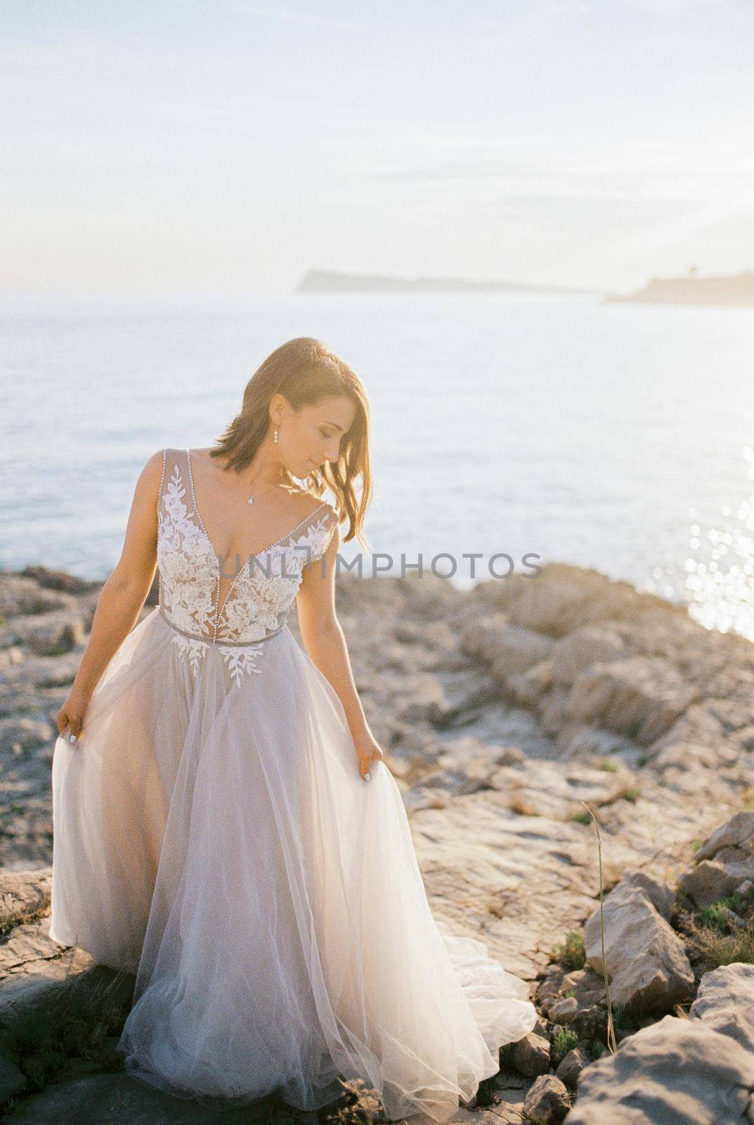 Bride in a white puffy dress stands on a rocky seashore holding her hem with her hands. High quality photo