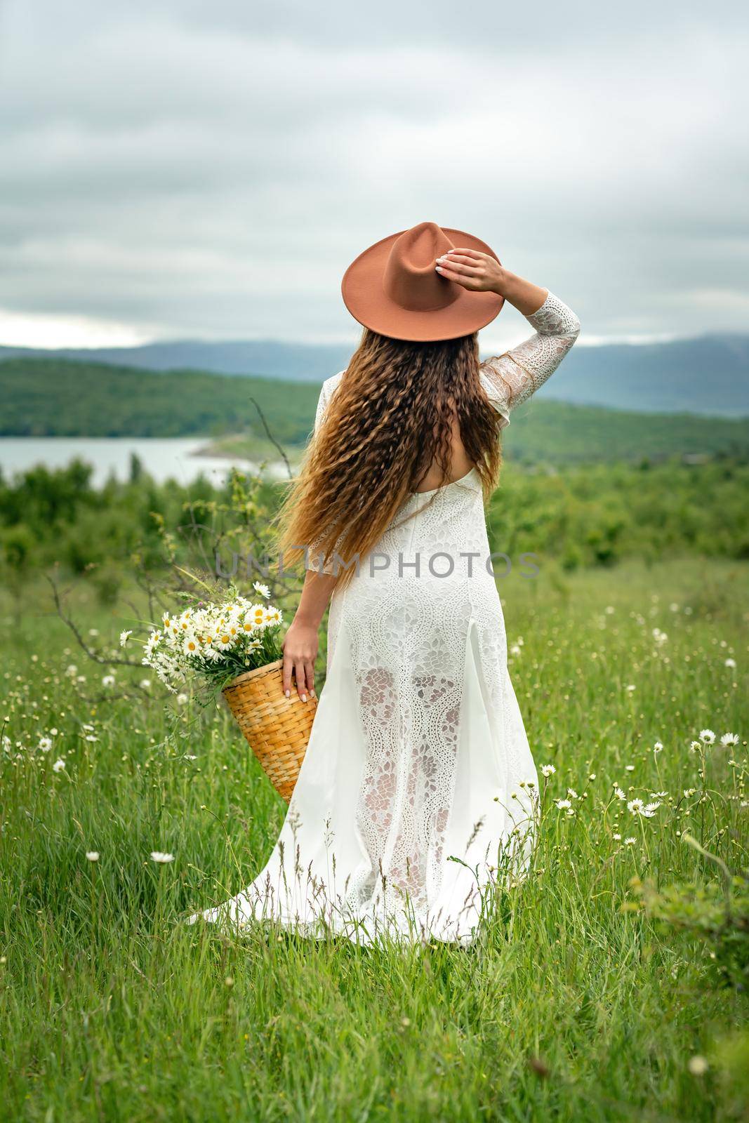 A middle-aged woman in a white dress and brown hat stands with her back on a green field and holds a basket in her hands with a large bouquet of daisies. In the background there are mountains and a lake