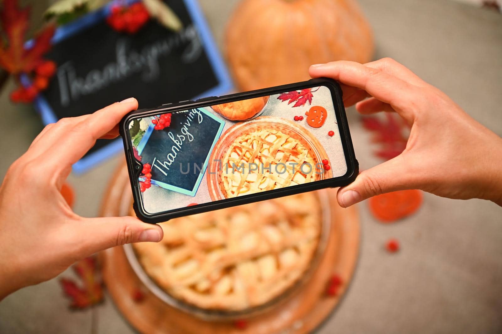 Woman's hands holding smartphone, taking photo of a freshly baked pumpkin apple pie with crusty lattice for a food blog by artgf