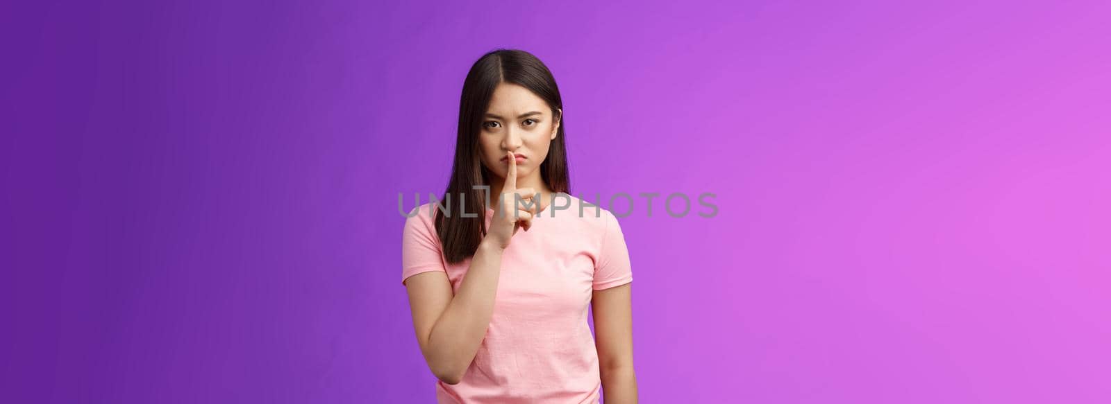 Shut up, seriously. Angry unsatisfied asian woman brunette frowning make hush sign, hold index finger on lips grimacing annoyed, prohibit speak, shushing stay silent, stand purple background.
