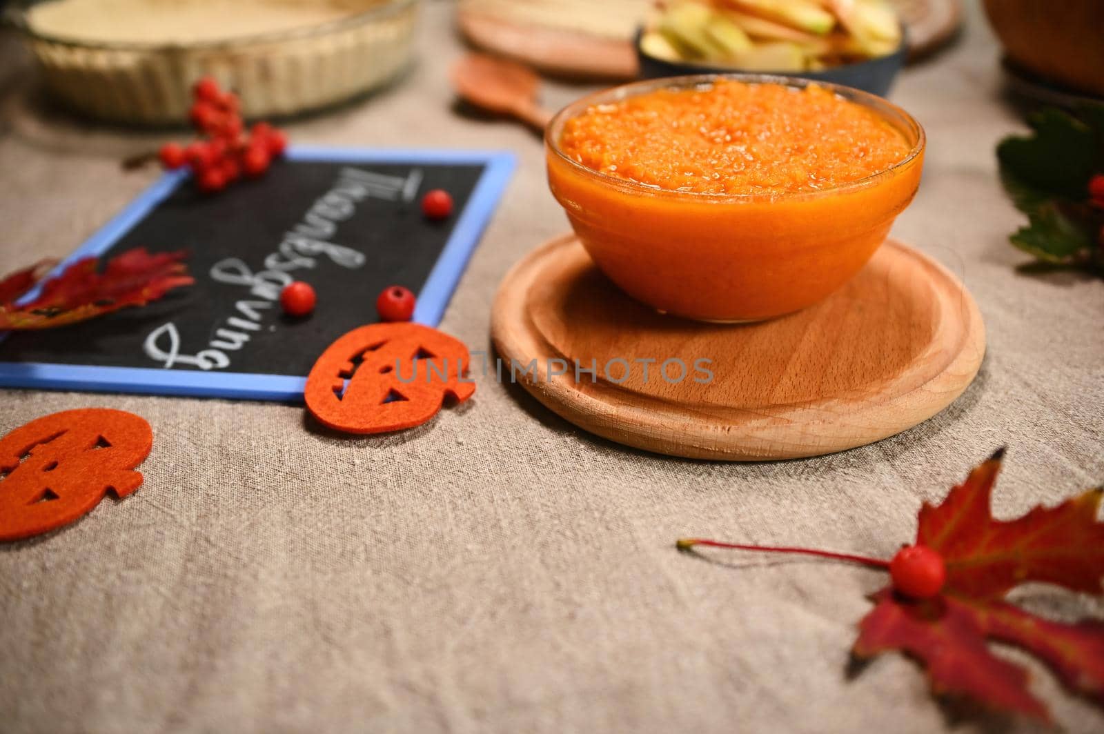 Glass bowl with mashed pumpkin, American classic pie with flaky crust, red autumn maple leaves on a chalkboard with lettering Thanksgiving, on a table with linen tablecloth. Autumnal holidays concept