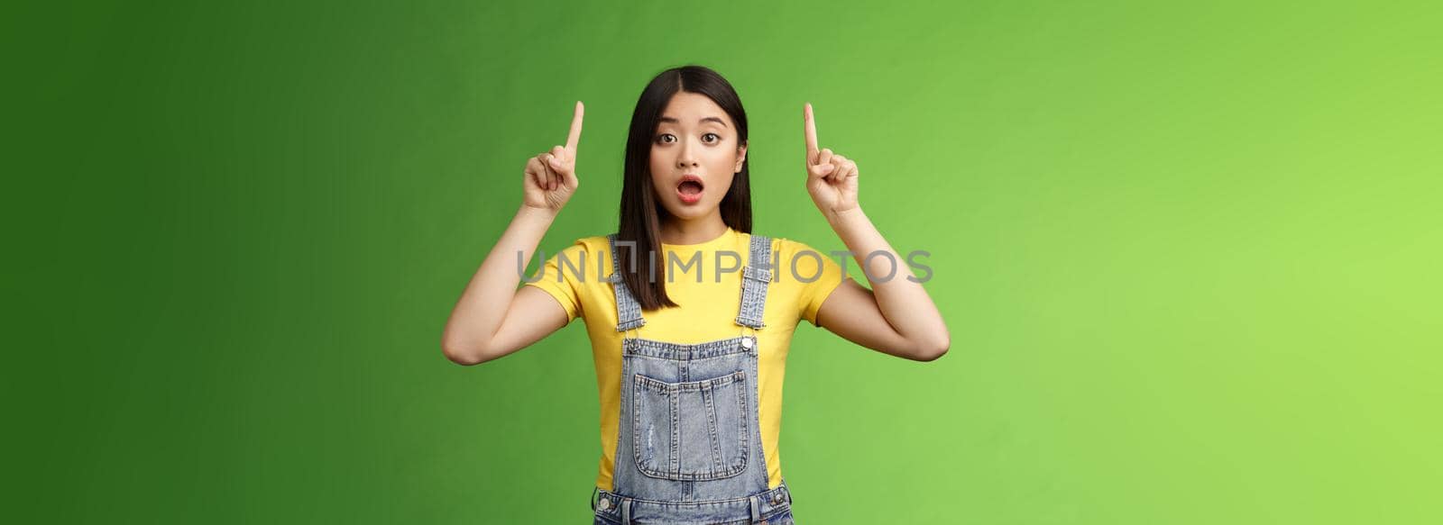 Impressed speechless shocked asian girl, pointing fingers up, drop jaw gasping astonished, stare camera amazed, telling about incredible prices new products, stand green background. Copy space