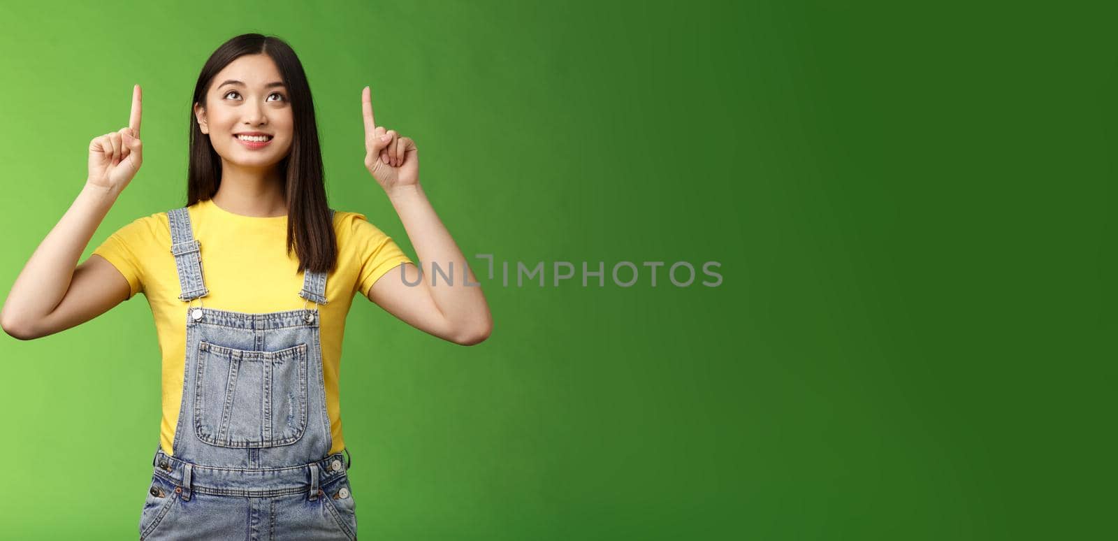 Dreamy hopeful cute asian girl look pointing up amused, smiling delighted, contemplate interesting object, enjoy stargazing, grinning satisfied, gazing top promo, stand green background.