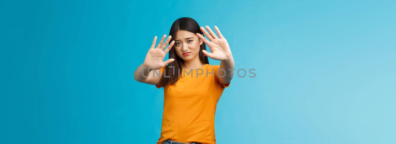 Asian moody displeased silly timid girl asking turn off light not photograph her, raise hands block, cover face step back and cringe disappointed, standing blue background upset reluctant. Copy space