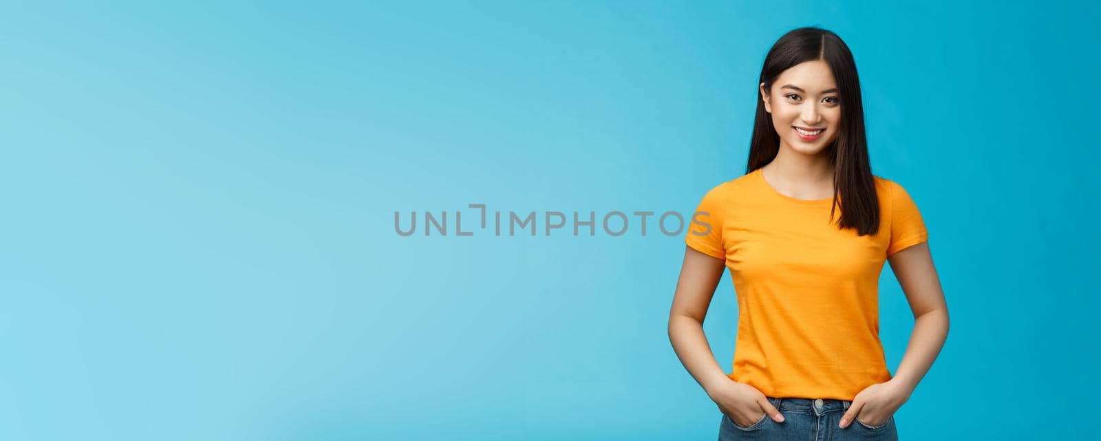 Confident assertive smiling asian woman stand blue background look camera, hold hands jeans pockets express self-assured vibe, enjoy seeing positive results skincare procedures, wear yellow t-shirt.