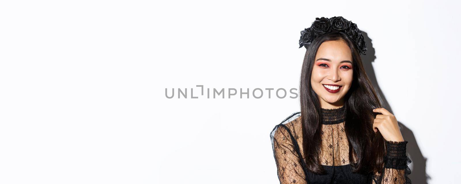 Close-up of beautiful elegant asian woman in black wreath and gothic lace dress smiling, standing over white background, dressed-up for halloween party.