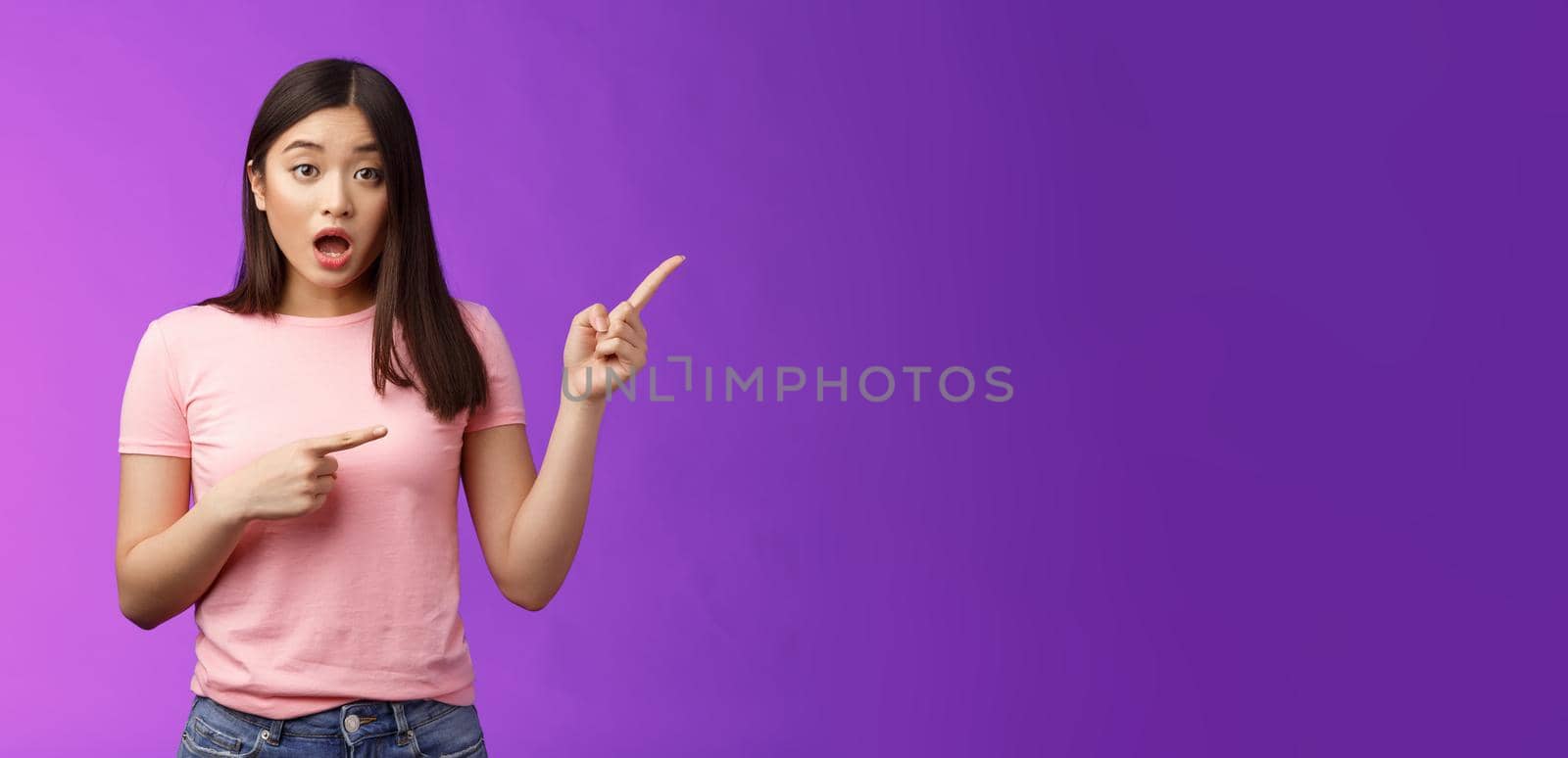 Excited alarmed cute asian female describe shocking scene pointing right, show index fingers sideways thrilling event, discuss amazing performance look camera worried, stand purple background.
