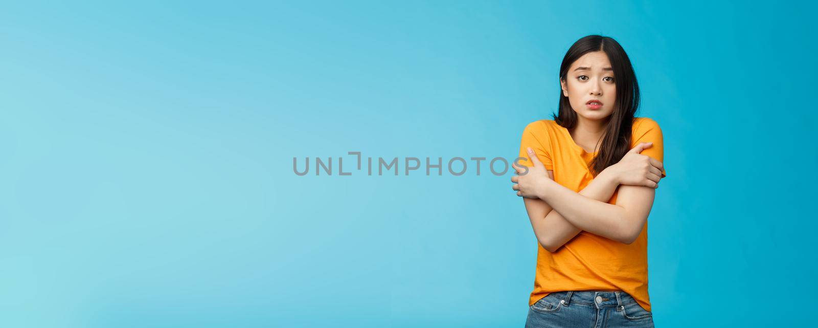Girl feeling uncomfortable walking light yellow t-shirt, hugging herself trembling, shaking feeling cold, freezing windy weather, frowning grimacing discomfort, stand blue background.