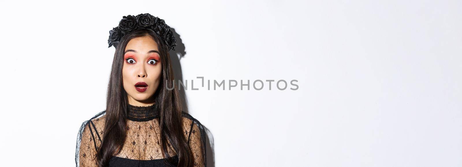 Close-up of impressed and shocked asian woman in halloween costume, drop jaw and gasping wondered, standing over white background.