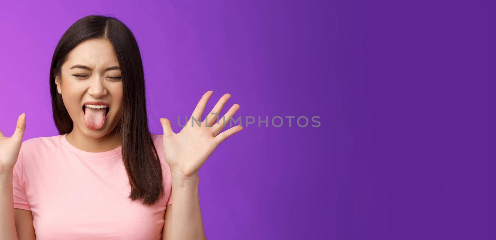 Close-up silly moody asian girl complaining act immature and bothered, raise hands unsatisfied show tongue aversion disgust, close eyes, react terrible disgusting thing, stand purple background.