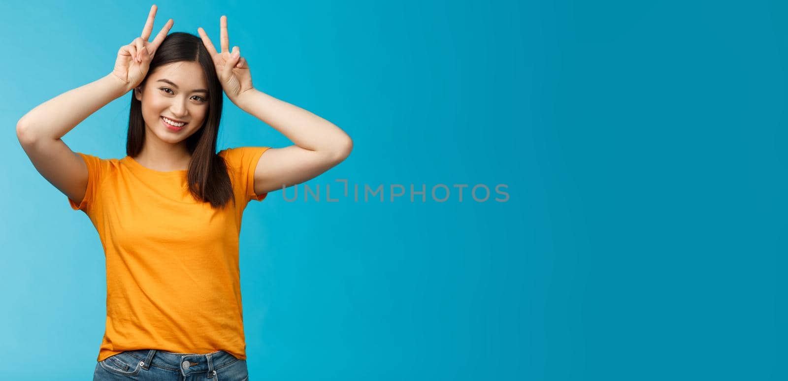 Cute tender asian girlfriend act silly lovely smile, tilt head show playfully ears hold peace victory signs on head, grinning toothy, express joy happiness and delight, stand blue background.