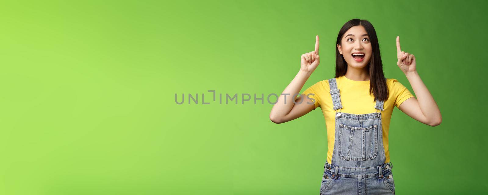 Carefree amused asian brunette in yellow t-shirt dungarees, smiling amazed and fascinated, look pointing up, observe cool new promo, grinning excited, enjoy watching top copy space.