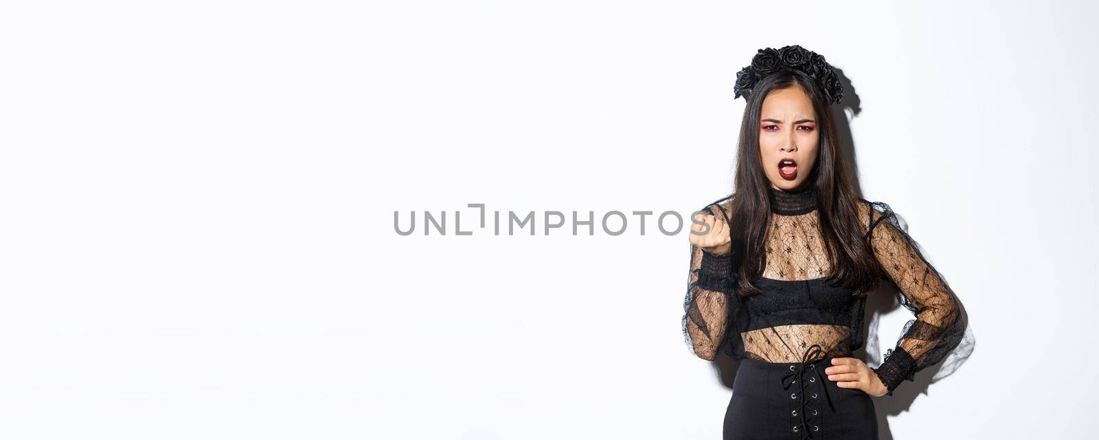 Image of angry and disappointed beautiful girl in halloween costume scolding someone. Girl shaking fist and looking with disapproval, wearing black gothic lace dress, standing white background.