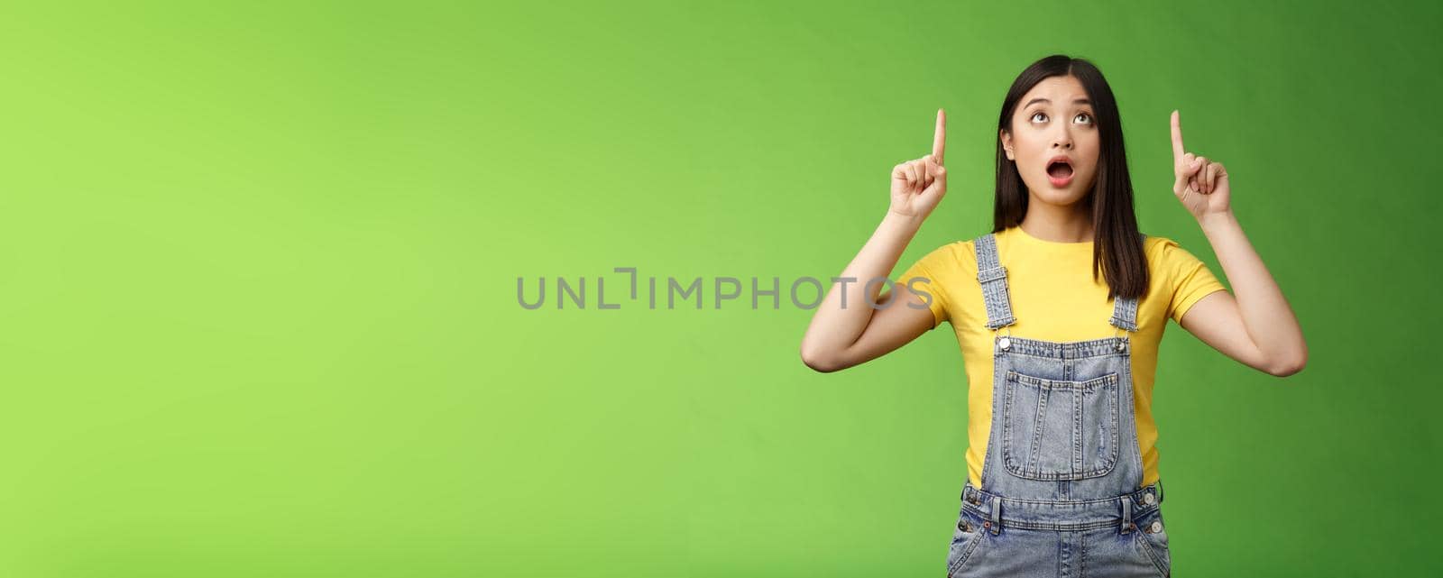 Omg look shocking price fall. Stunned speechless amazed asian girl drop jaw, wondering what happening upstairs, pointing lookin up impressed, staring surprised stand green background by Benzoix