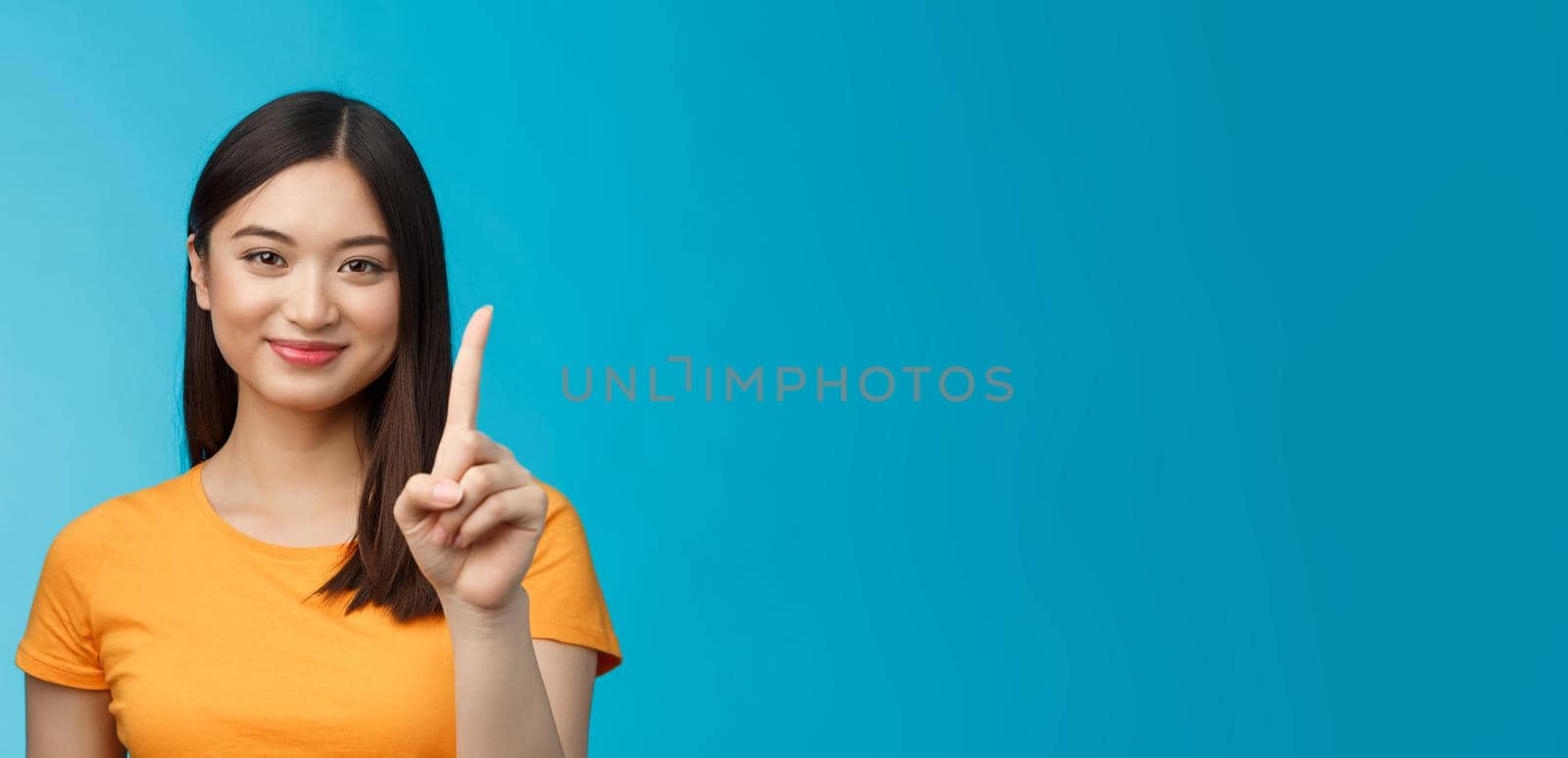 Close-up cheerful attractive tender asian woman with dark short haircut show number one, extend forefinger, smiling joyfully, friendly grin, stand blue background relaxed, casually make order.