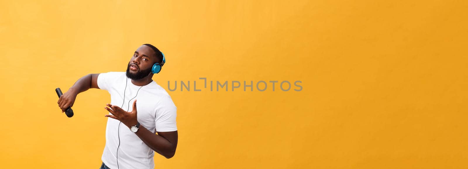 Portrait of cheerful positive chic. handsome african man holding microphone and having headphones on head listening music singing song enjoying weekend vacation isolated on yellow background.