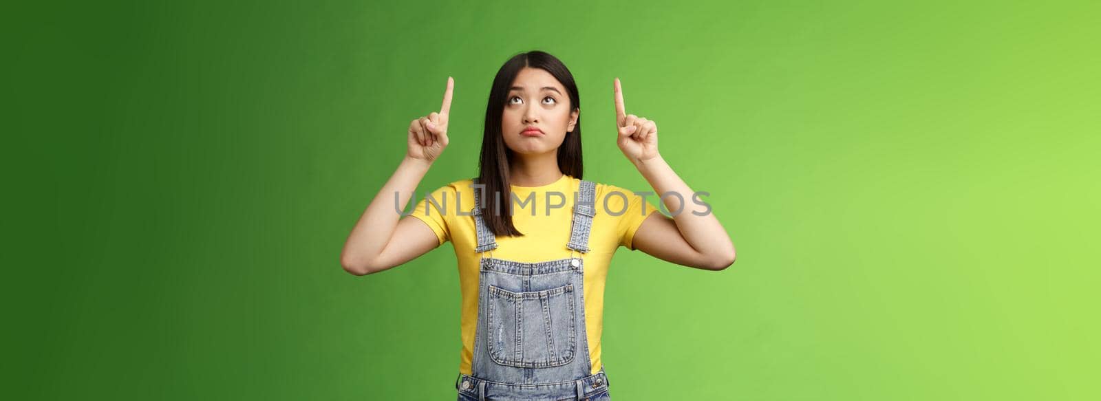 Silly upset cute asian girl look pointing up sad, gloomy watching top side, grimacing displeased, dreaming have new product, regret not have money effort cool thing, stand green background by Benzoix