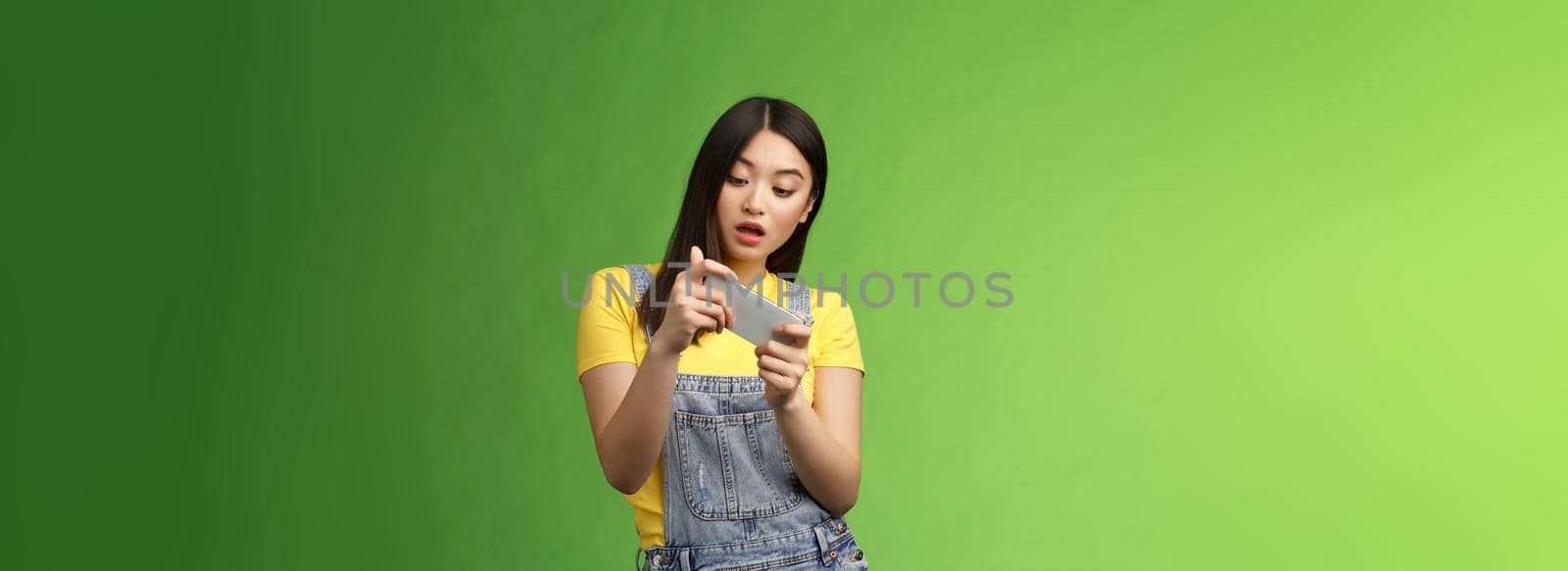 Carefree geeky cute asian brunette waste time university campus, playing online smartphone game, look amused entertained telephone screen, tilting body move character, open mouth excited by Benzoix