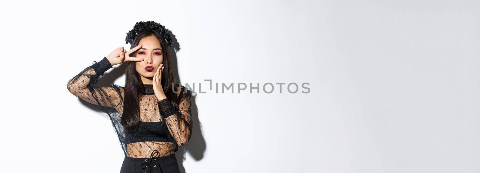 Image of beautiful asian woman in gothic lace dress and black wreath posing on halloween banner, showing peace gesture and pouting, standing over white background.