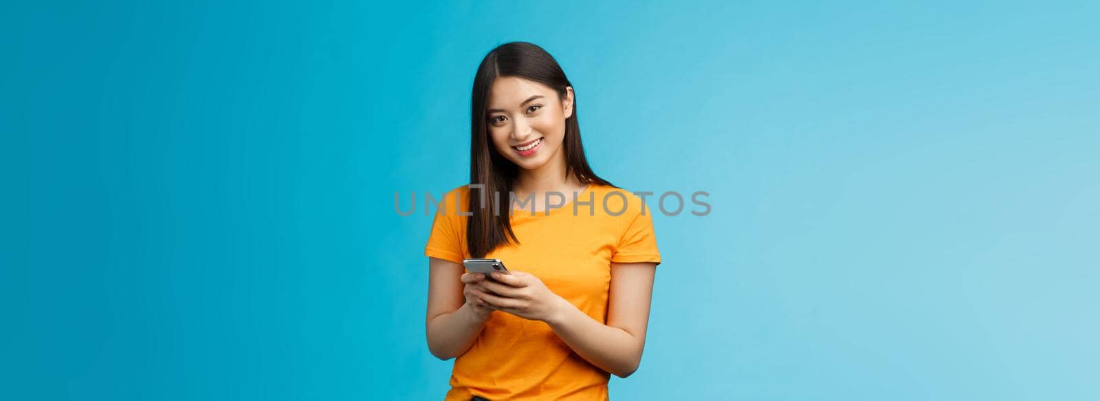 Cheerful friendly modern young asian girl student messaging friend, hold smartphone texting followers, using new internet application, try out app stand satisfied smiling camera, blue background by Benzoix
