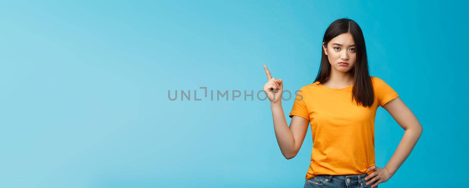 Moody upset asian girlfriend, sulking look offended and displeased pointing upper left corner, pouting disappointed, standing distressed blue background wear yellow t-shirt. Copy space