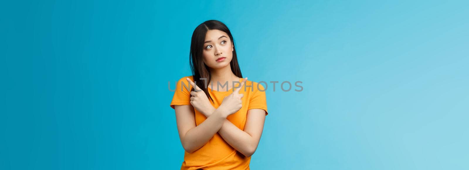 Confused cute asian female student thinking what do, look sideways thoughtful, cross hands chest pointing left and right, making choice, thinking what pick, taking important decision blue background.